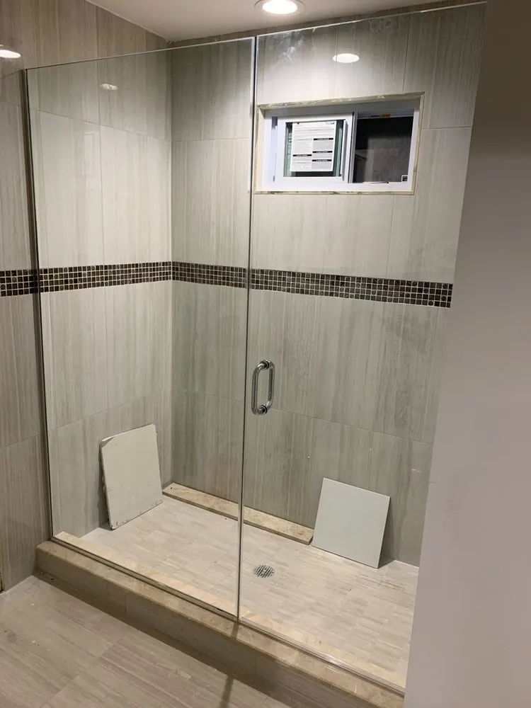 Glass shower with gray linear wall tile, brown glass mosaic band and a window, loose tile pieces still on the floor.