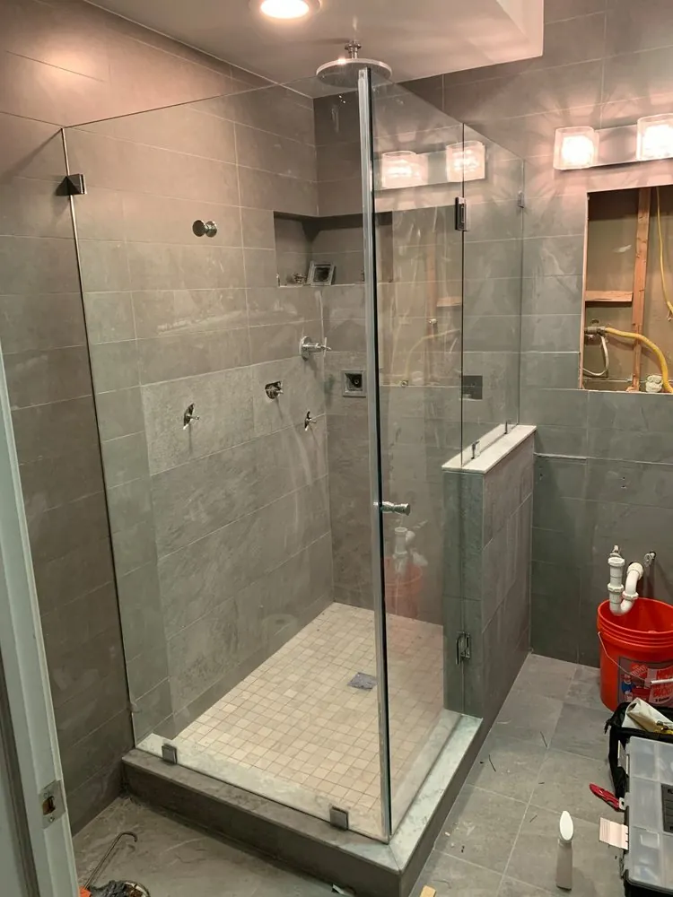 Bathroom mid-renovation with a new gray tile shower and glass enclosure, open framed wall, exposed plumbing and tools.