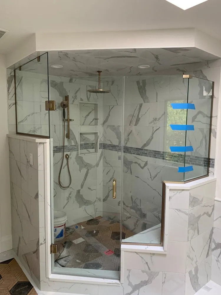 Neo-angle glass shower with gold fixtures, white marble-look tile, mosaic band and niches, bucket and debris inside.