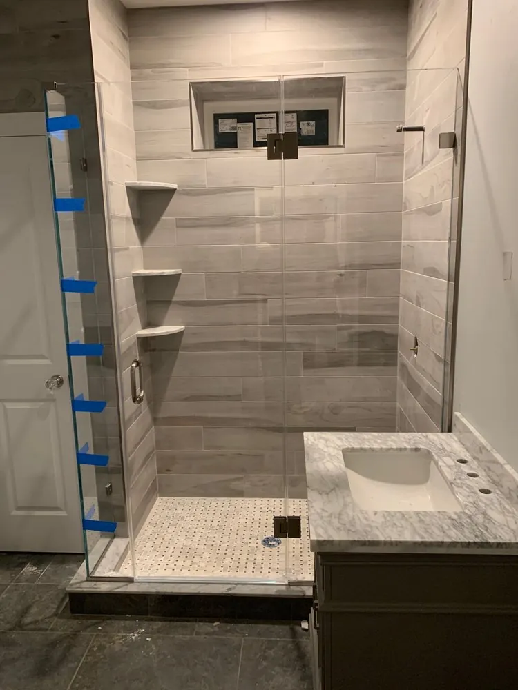 Shower with gray wood-look plank tile, basketweave floor and corner shelves beside a marble-top vanity, nearly done.
