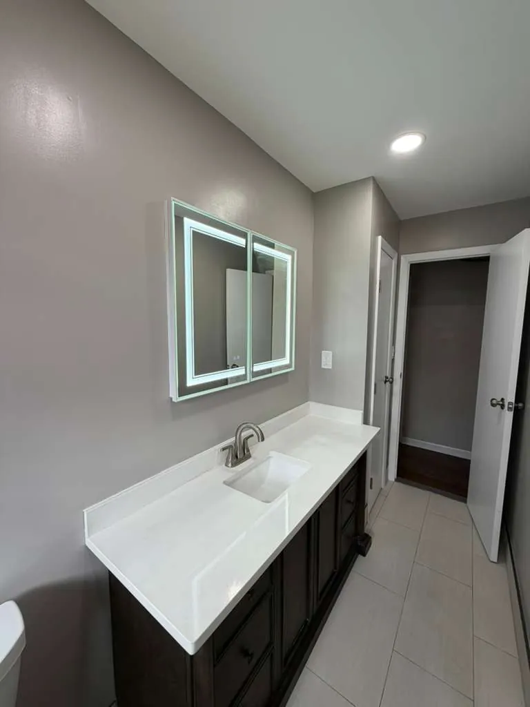 Espresso vanity with white countertop and brushed nickel faucet below an LED mirror cabinet on a gray wall.