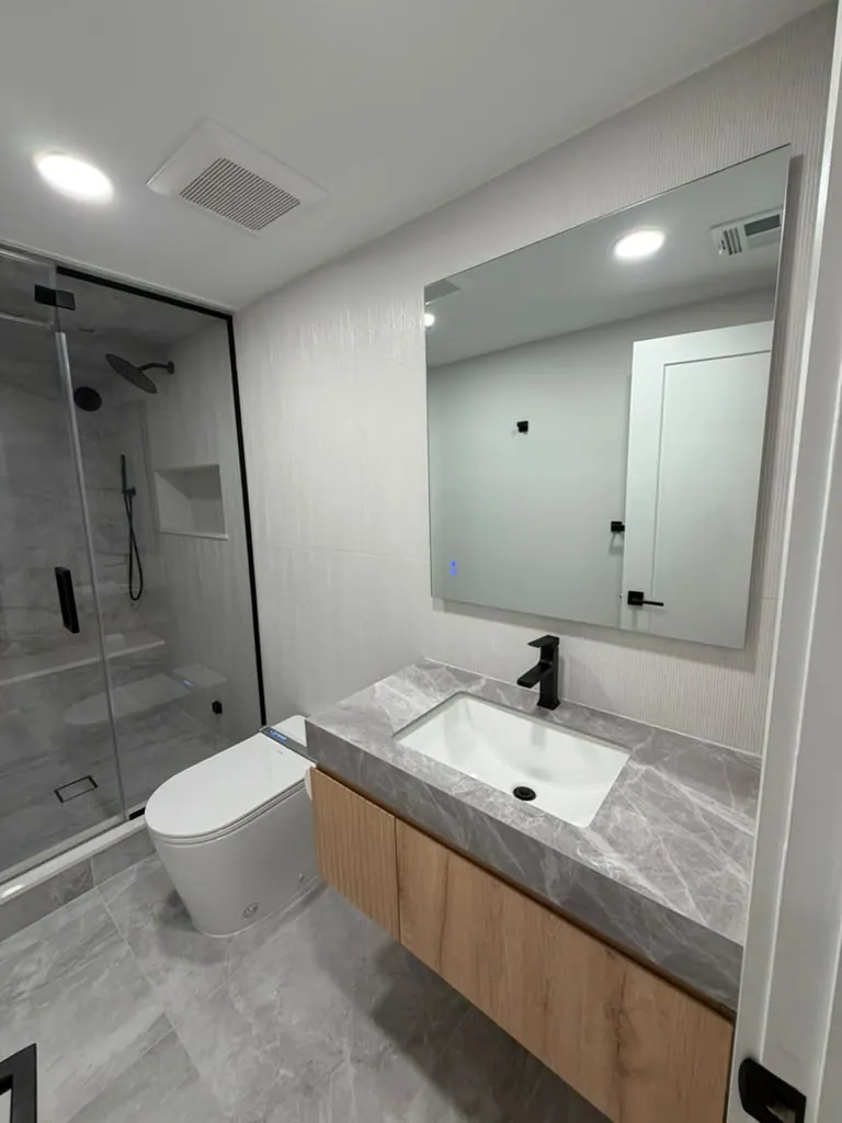 Bathroom with a floating wood vanity, thick gray stone top, black faucet, large mirror and a glass shower beside the toilet.