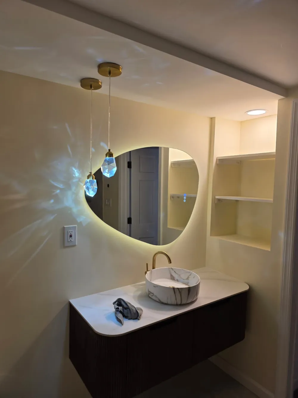 Floating fluted wood vanity with a marble-look vessel sink, gold faucet, backlit pebble mirror and crystal pendant lights.