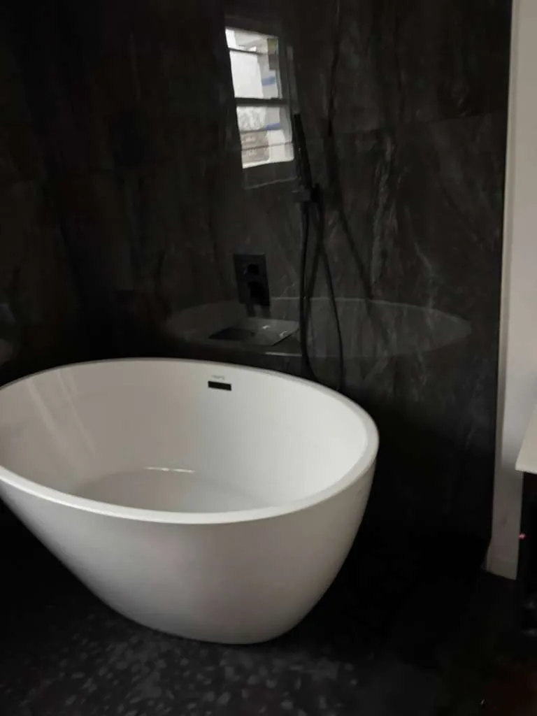 White oval freestanding tub against dark marble-look wall panels with a black handheld shower and pebble floor.