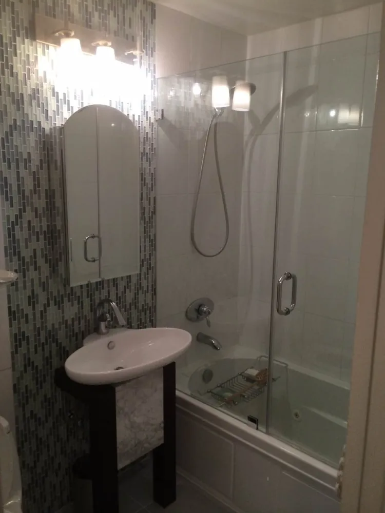Bathroom with blue-green glass mosaic wall, oval vessel sink on dark stand, arched mirror and tub with hinged glass door.