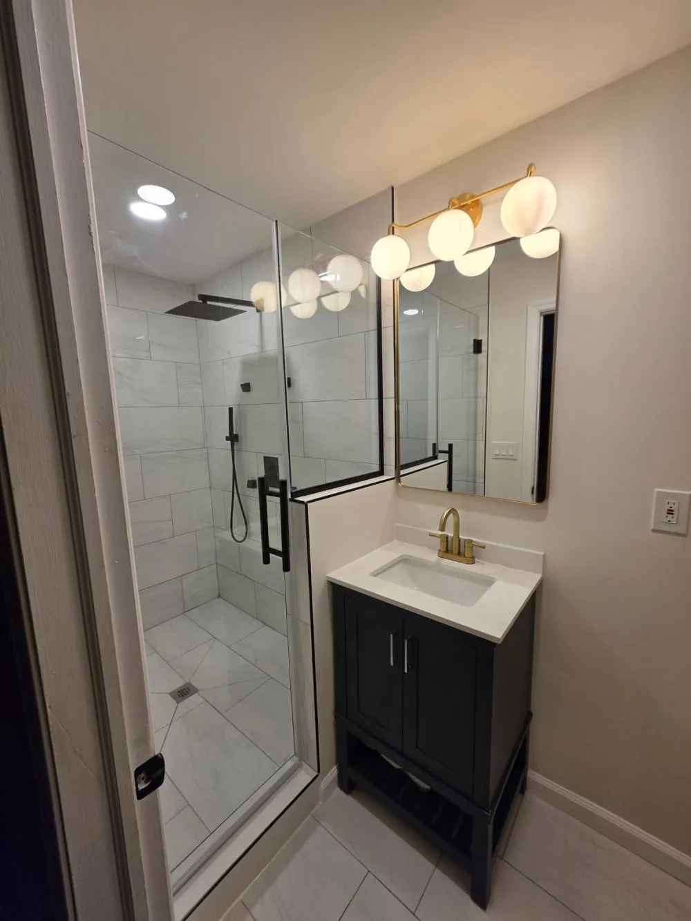 Bathroom with glass walk-in shower in white tile, black vanity with brass faucet, gold mirror and globe vanity light.