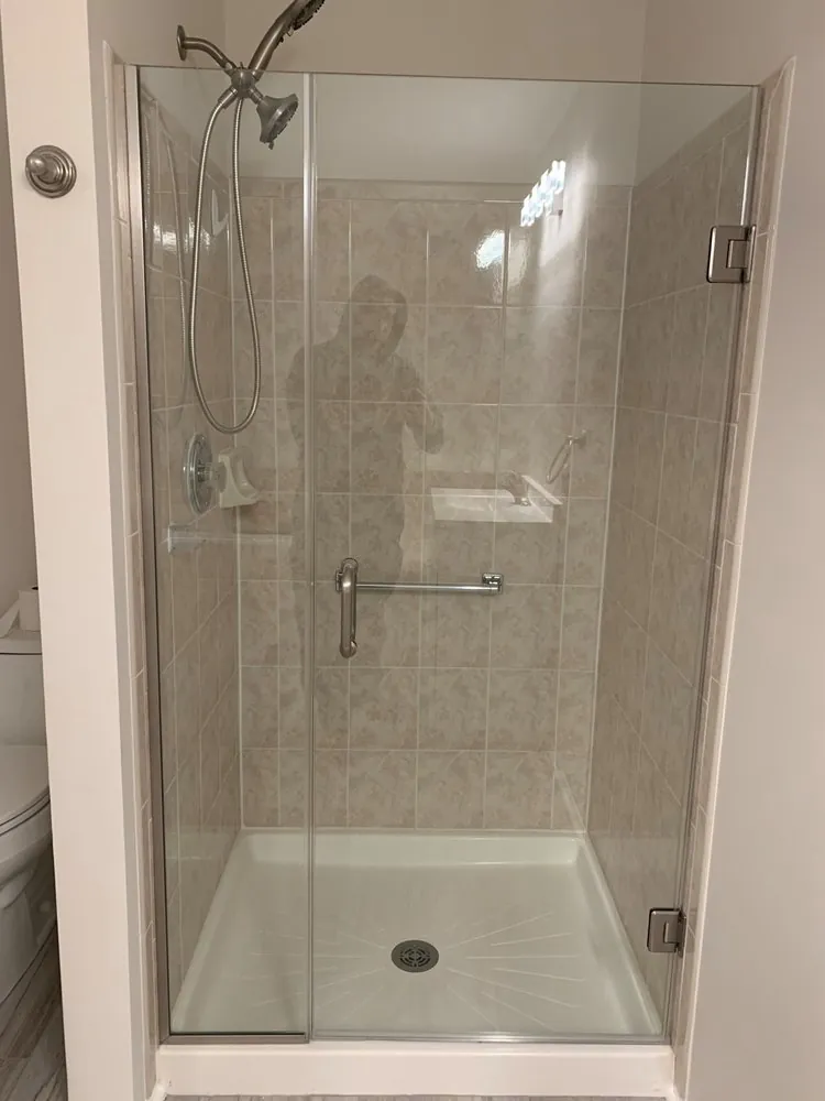 Frameless hinged glass shower door with towel bar over a white shower base with beige tile walls.