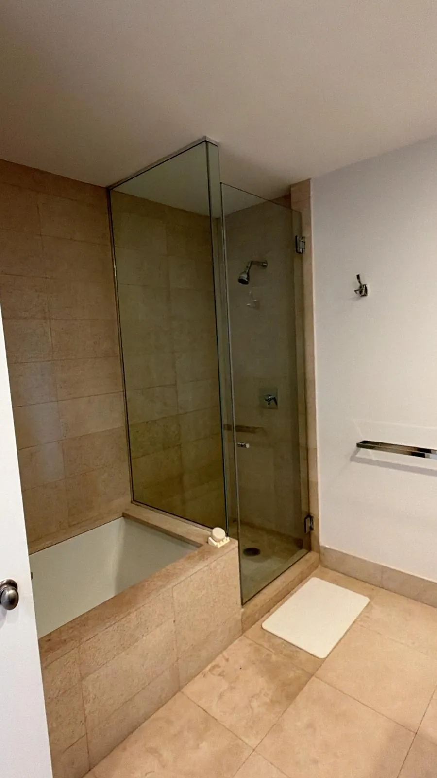 Corner glass shower enclosure next to a stone-clad soaking tub, with beige stone tile floor and walls.