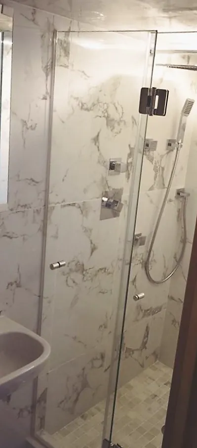 Narrow glass shower with white marble-look porcelain walls, chrome hand shower and valves, small mosaic floor.