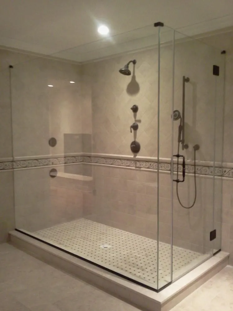 Large glass shower with beige travertine tile, scroll border, basketweave floor and bronze fixtures.