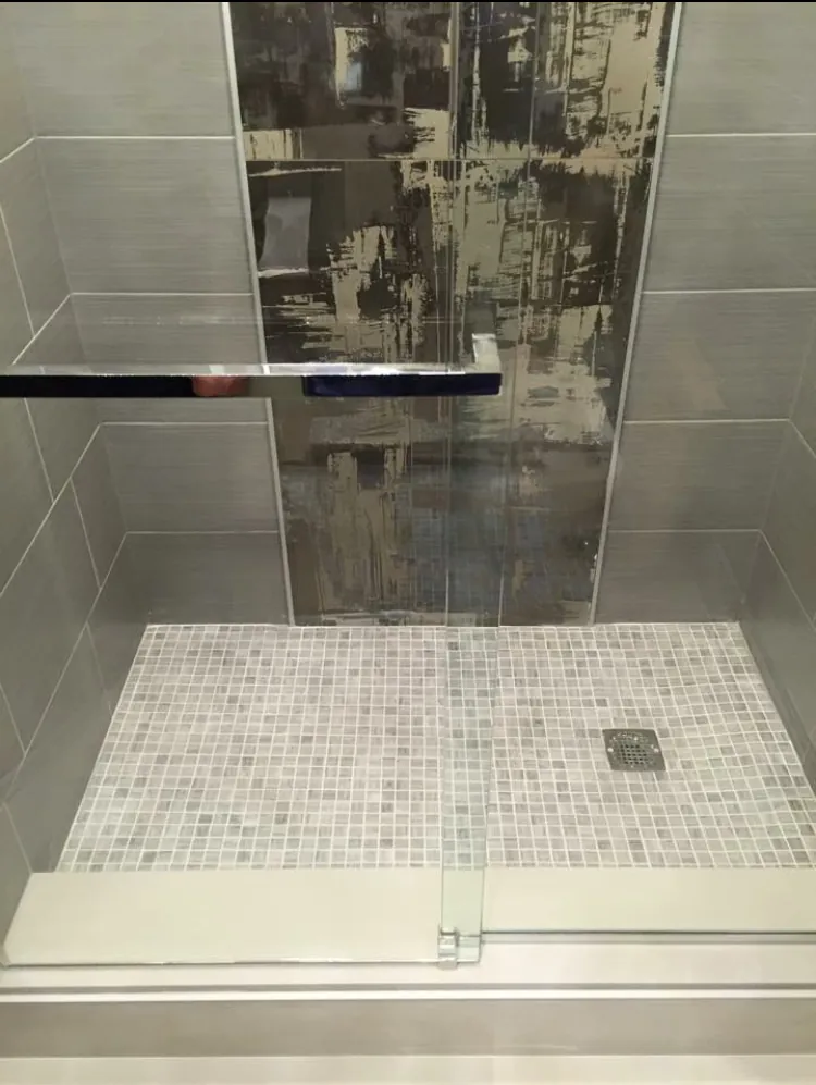 Shower floor with gray square mosaic tile below gray wall tile and a black and gold abstract accent panel.