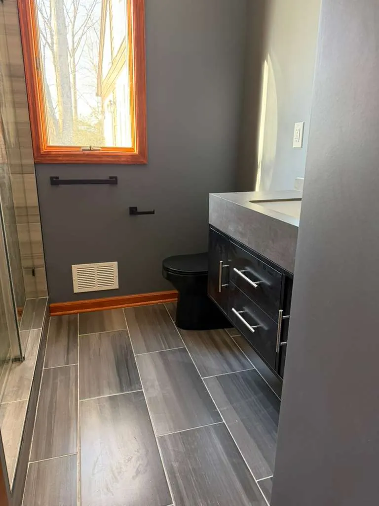 Bathroom with gray wood-look plank tile floor, black toilet, dark gloss vanity with gray top and black towel bar.
