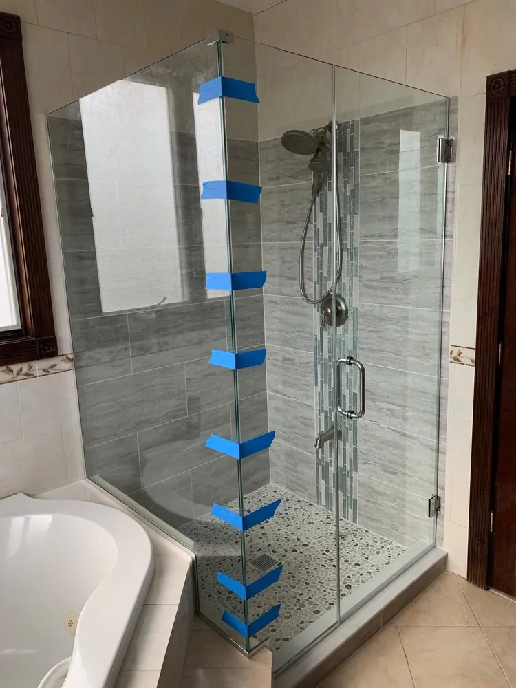 Frameless glass shower with gray plank tile, vertical glass mosaic strip, pebble floor, beside a corner jetted tub.