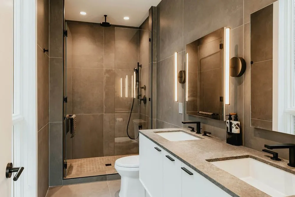 Bathroom with large gray porcelain tile, walk-in glass shower, white double vanity, stone top, black faucets and light bars.