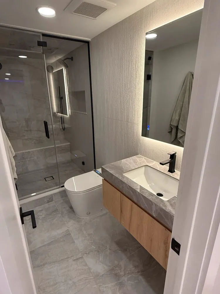 Modern bathroom with gray stone-look tile, black-framed glass shower, floating wood vanity, backlit mirror and black faucet.