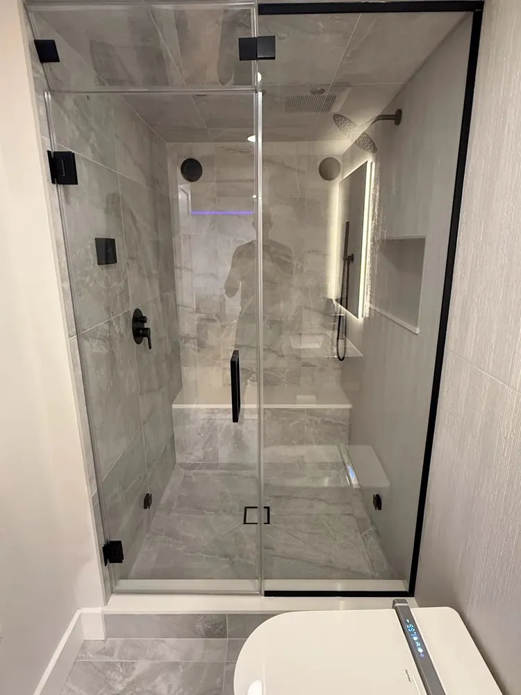 Black-framed glass shower with gray stone-look tile, built-in bench, niche, lighted mirror and matte black fixtures.