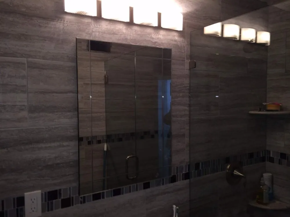 Bathroom wall of gray vein-cut tile with glass mosaic border, frameless mirror, vanity light bar and glass shower panel.