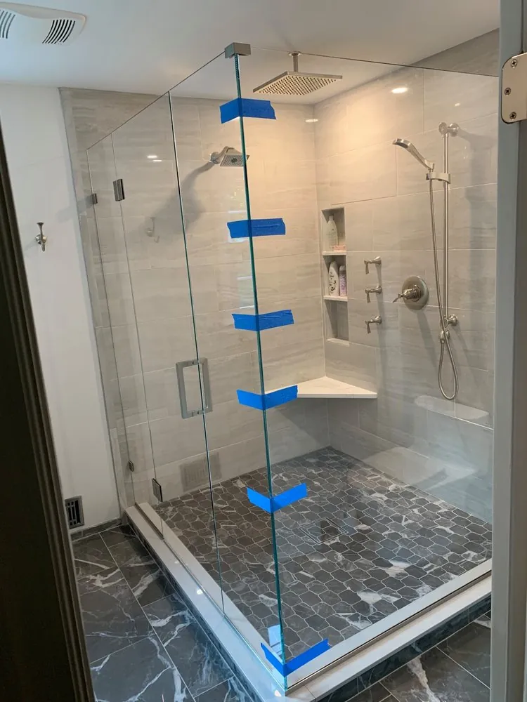 Frameless glass shower with gray plank tile, rain and handheld heads, corner bench, niche and dark marble mosaic floor.