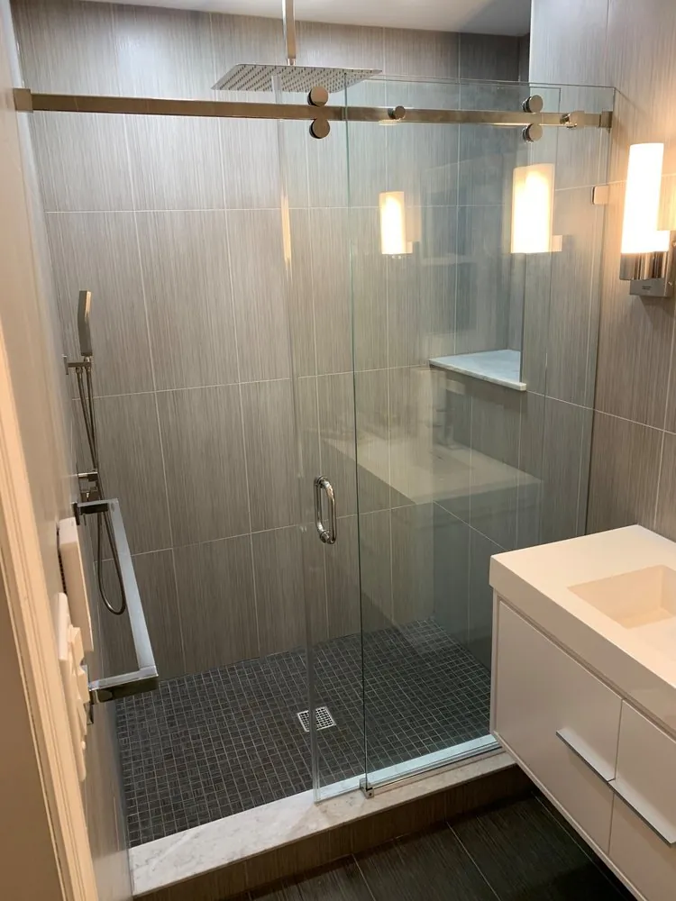 Glass shower with gray linear tile, rain head, dark mosaic floor and marble curb beside a white floating vanity.