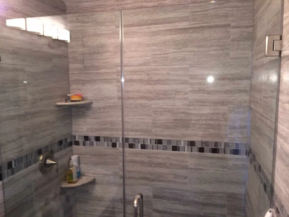 Shower with gray vein-cut wall tile, glass mosaic border, corner shelves, brushed nickel valve and frameless glass door.