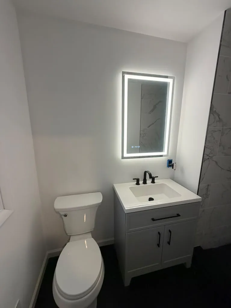 Bathroom with a gray vanity, white sink top, black faucet and pulls, lighted LED mirror and dark floor tile.