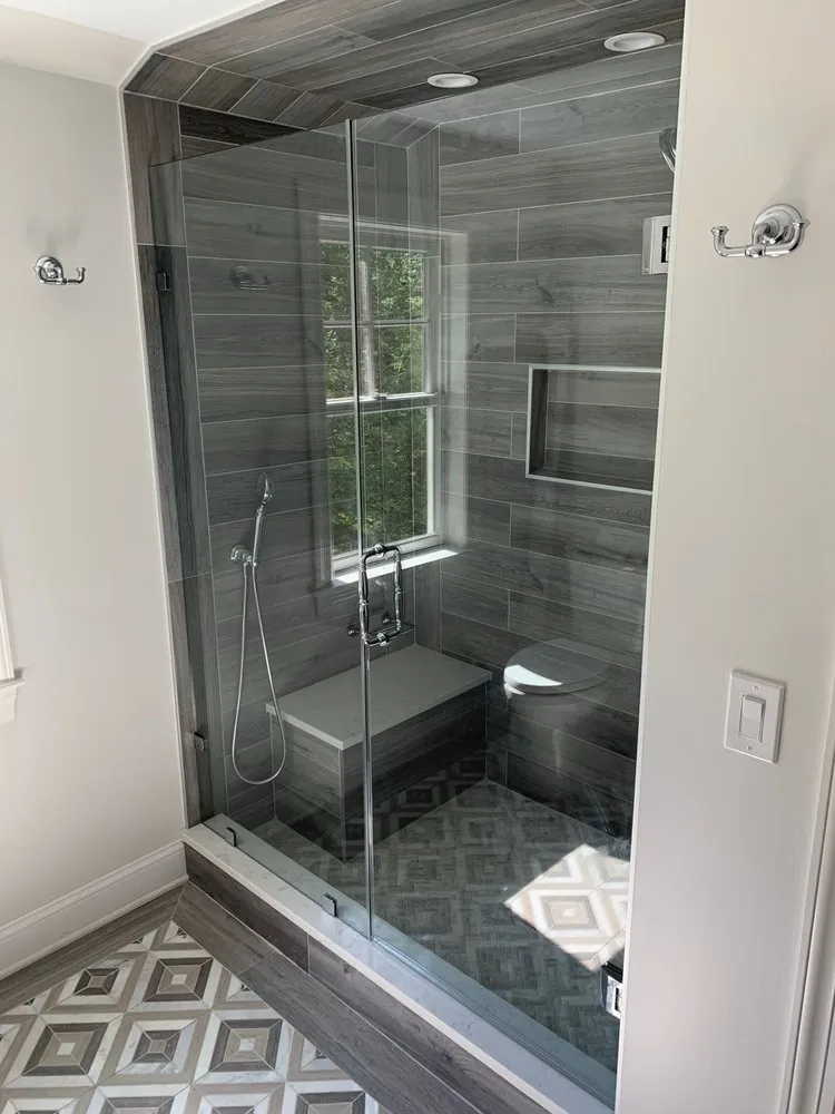 Glass shower with gray wood-look plank tile, window, quartz bench, niche and patterned floor tile outside the shower.