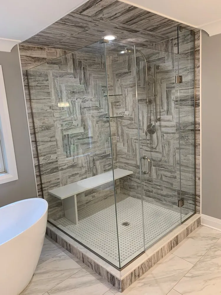 Frameless glass shower with gray wood-look herringbone tile on walls and ceiling, white floating bench and basketweave floor.