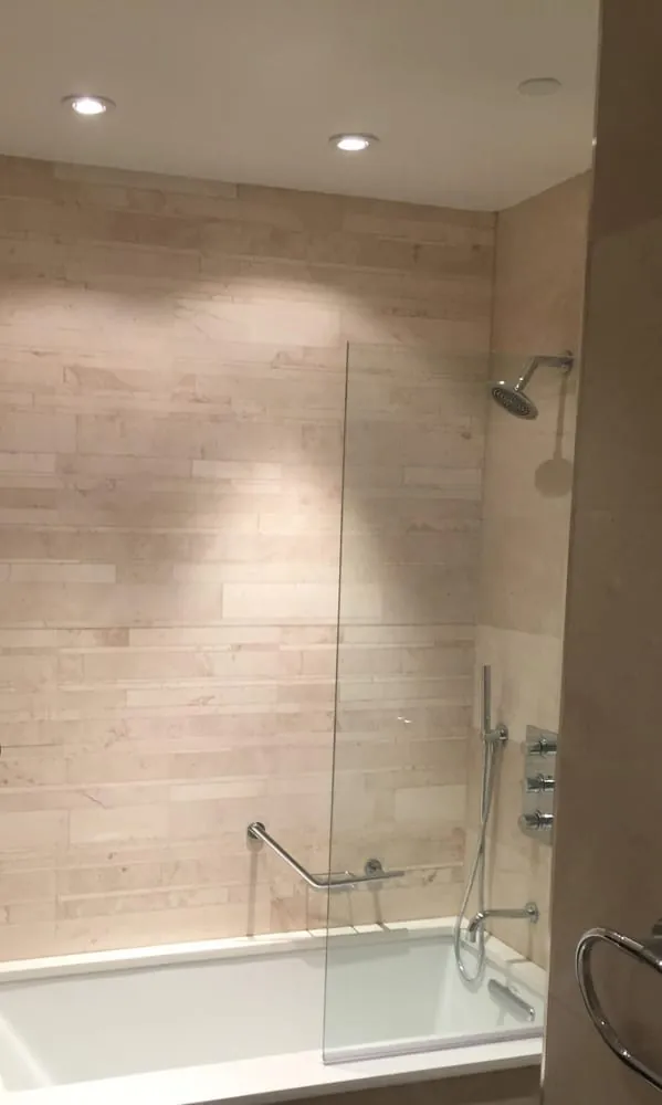 Soaking tub with cream limestone ledger tile surround, glass splash panel and chrome shower fixtures.