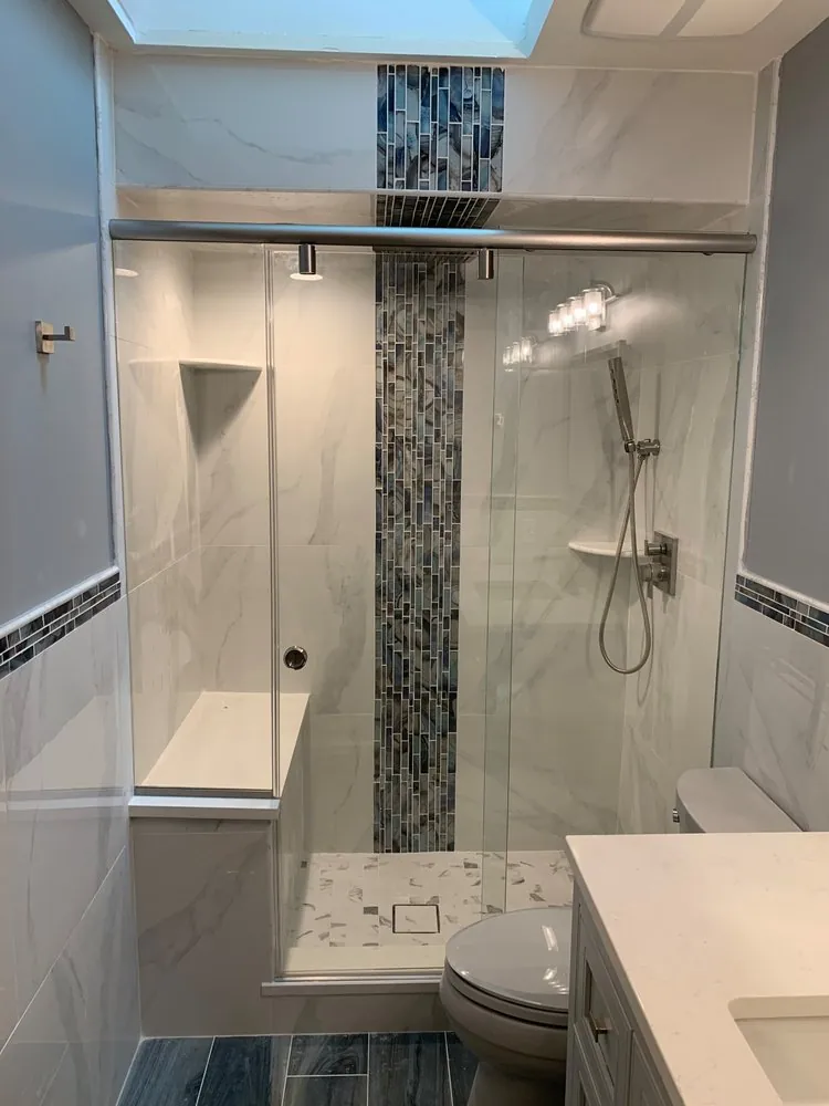 Shower with white marble-look tile, vertical blue glass mosaic stripe, bench, sliding glass door and skylight above.