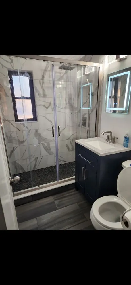 Small bathroom with marble-look shower walls, pebble shower floor, sliding glass doors, navy vanity and lighted mirror.