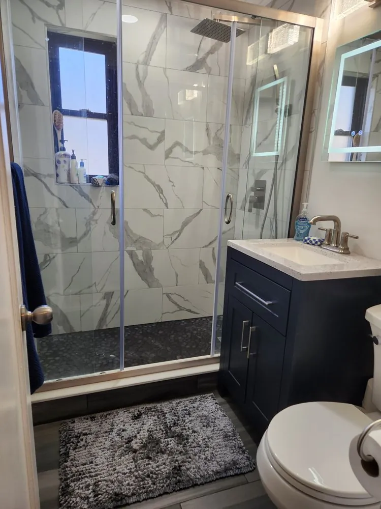 Walk-in shower with white marble-look tile, black pebble floor, sliding glass door and a navy vanity with quartz top.