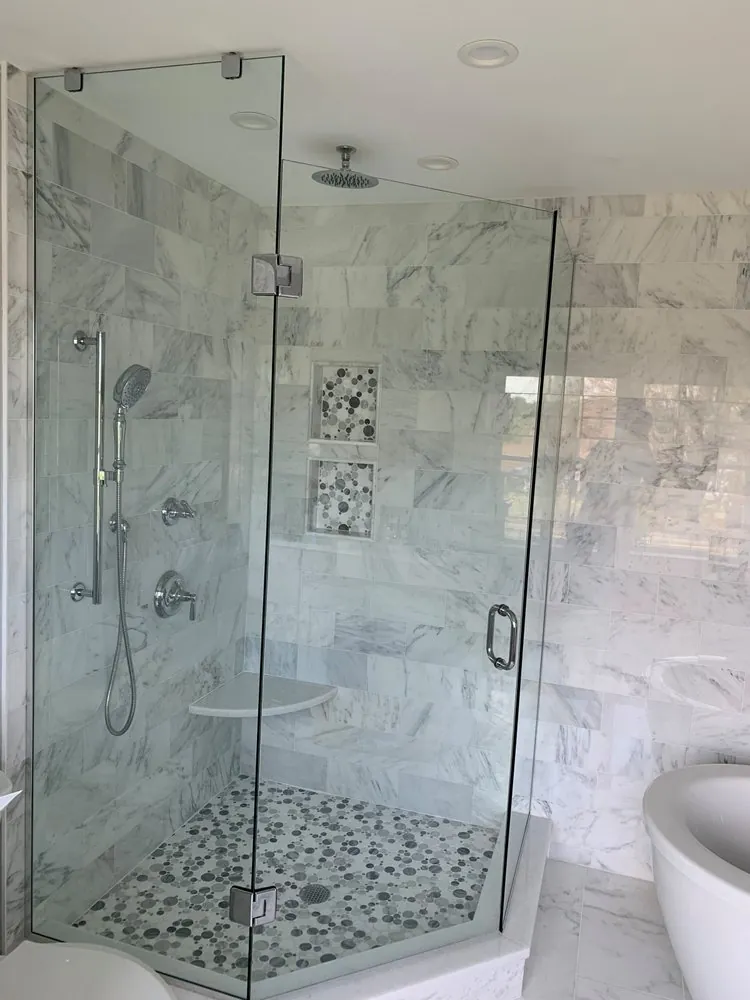 Neo-angle frameless glass shower with white marble tile, pebble mosaic floor and niches, ceiling rain head and corner seat.