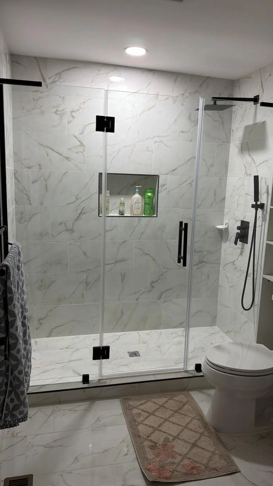 Walk-in shower with white marble-look tile, frameless glass door with black hinges, niche and black rain showerhead.
