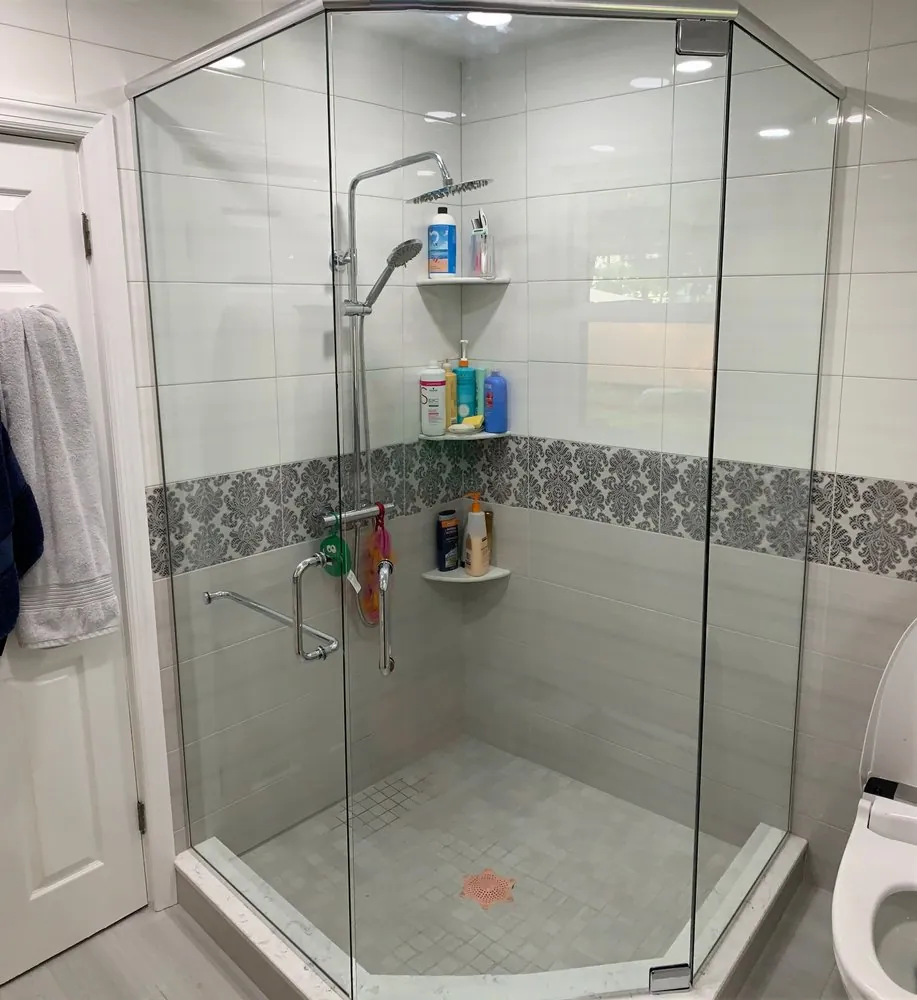 Neo-angle glass shower with white and gray tile, a damask-pattern border, corner shelves and gray mosaic floor.