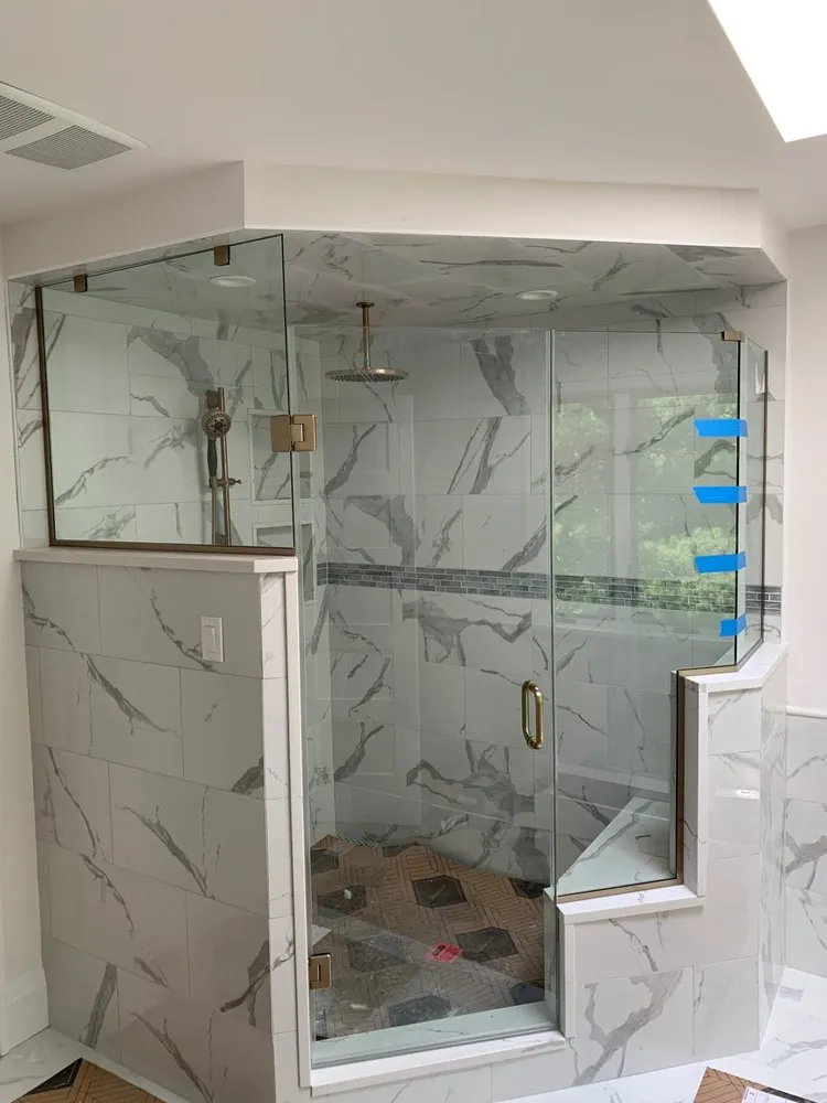 Neo-angle glass shower with brushed gold hardware, white marble-look tile, gray mosaic band and a tiled bench.