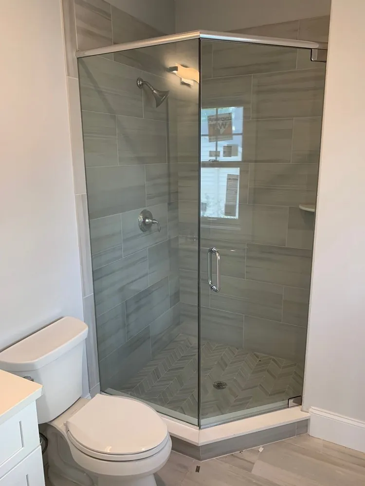 Neo-angle glass shower with gray linear stone-look tile and chevron floor next to a toilet and white vanity.