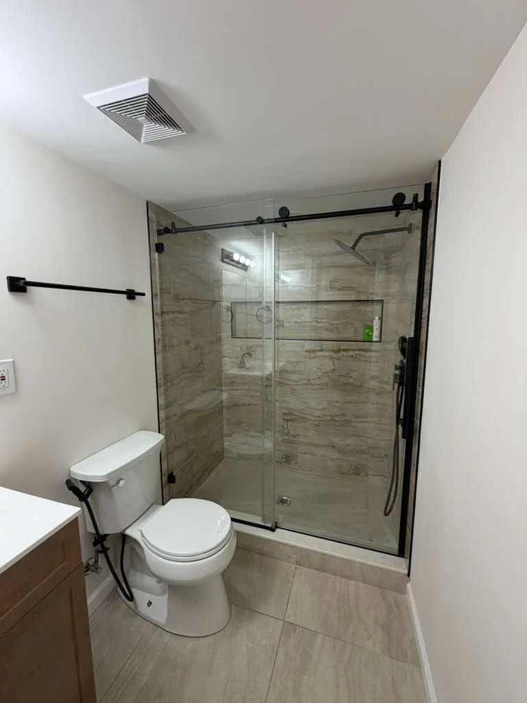 Bathroom with beige stone-look shower tile, long niche, black sliding glass door, white toilet and wood vanity.