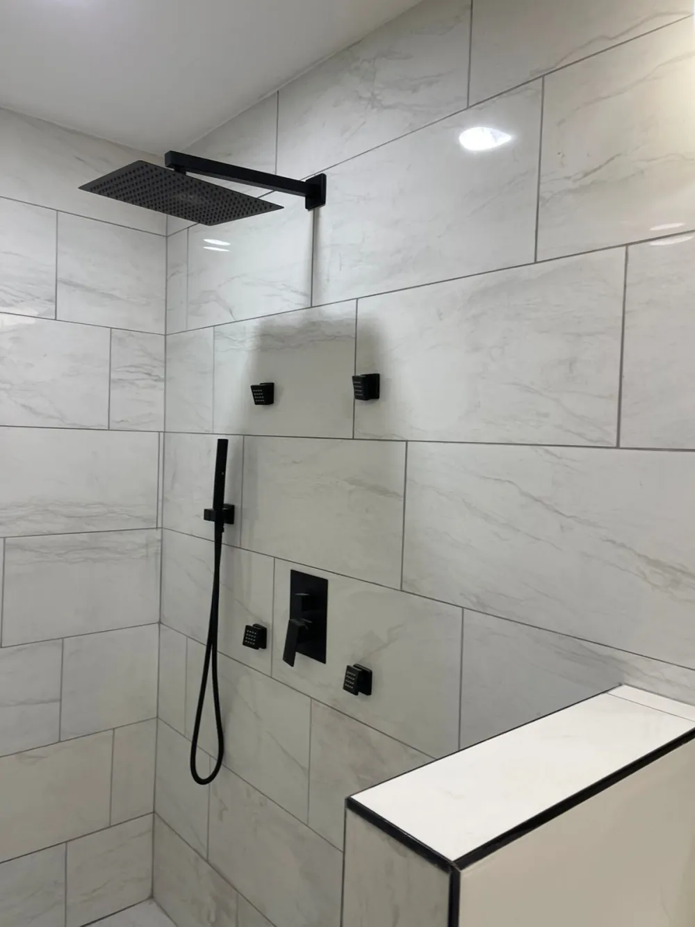 Shower with large white marble-look tile, black rain showerhead, handheld wand, four body jets and a tiled half wall.