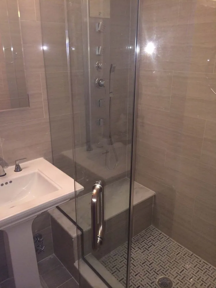 Glass shower with taupe linear tile, body spray jets, handheld shower, marble bench, basketweave floor and pedestal sink.