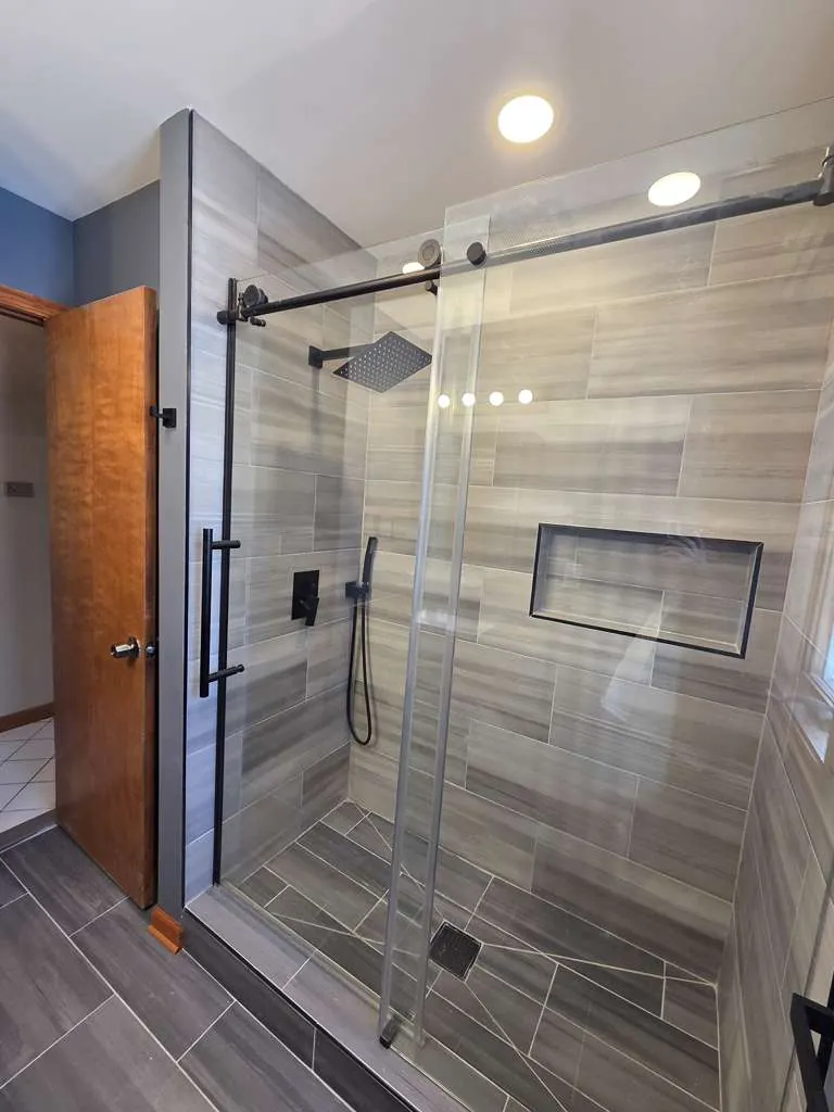 Walk-in shower with gray wood-look plank tile, long niche, rain head and black sliding glass door hardware.