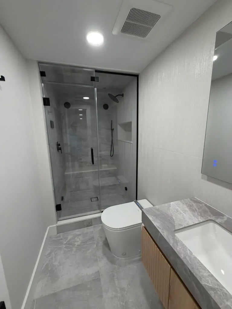 Gray tile shower with frameless glass, bench, niche and black fixtures, next to a smart toilet and gray stone vanity top.