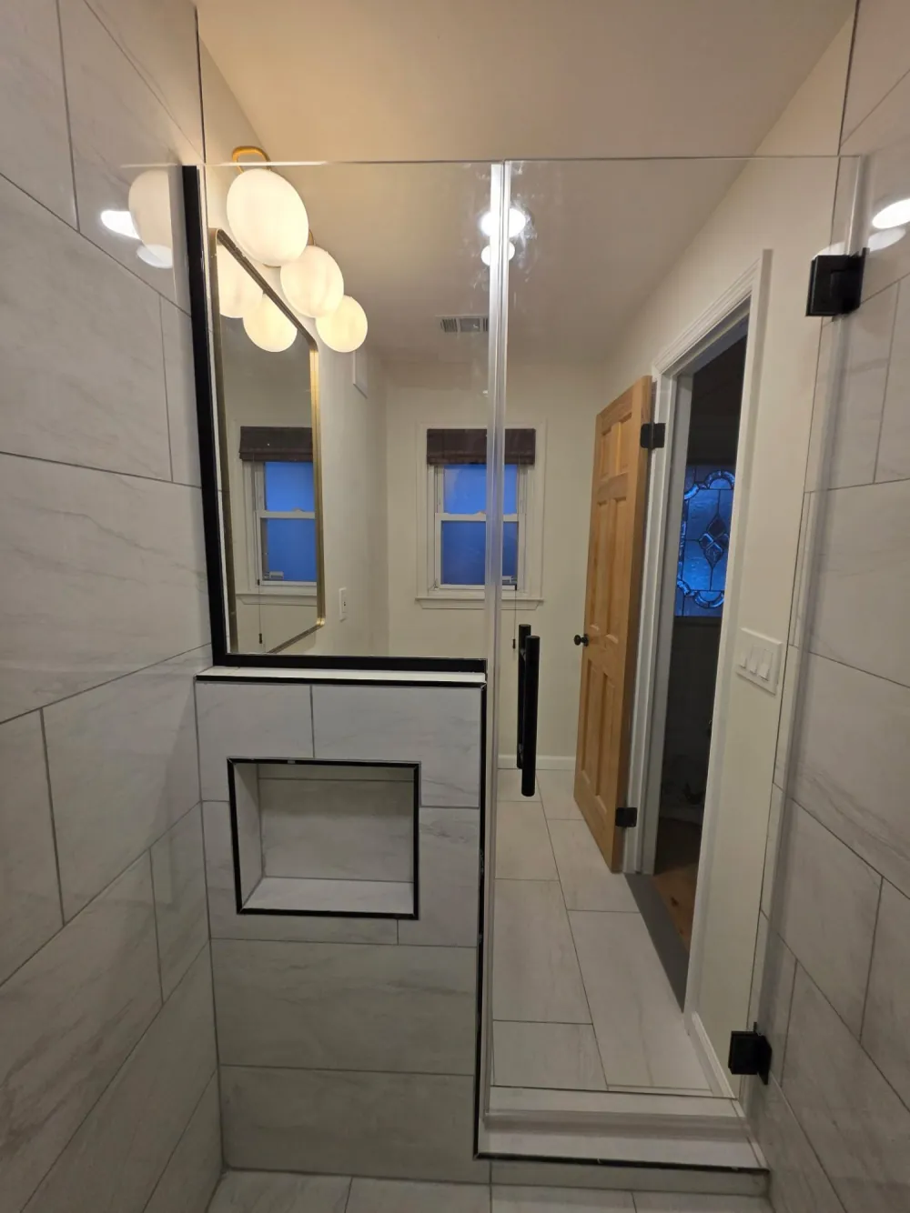 View from inside a shower: tiled half wall with black-trimmed niche, glass panel and door, white plank tile floor.