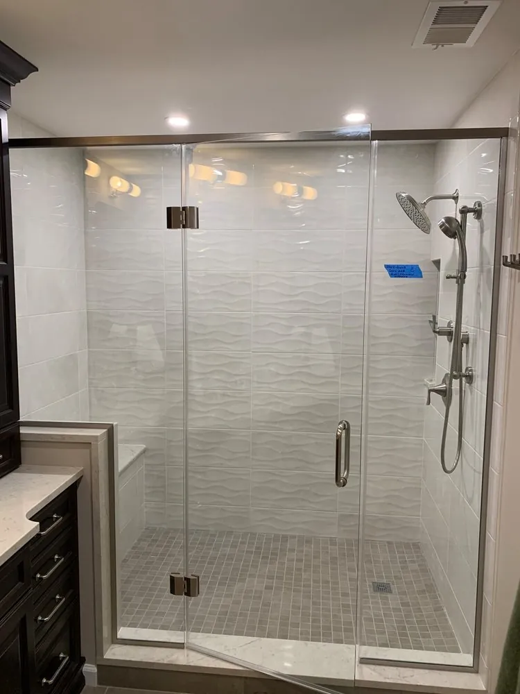 Frameless glass shower with white wave-textured wall tile, gray mosaic floor, brushed nickel fixtures and a dark vanity.