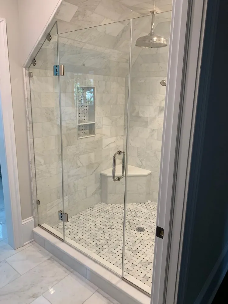 Frameless shower under a sloped ceiling with white marble subway tile, basketweave floor, corner bench and rain head.