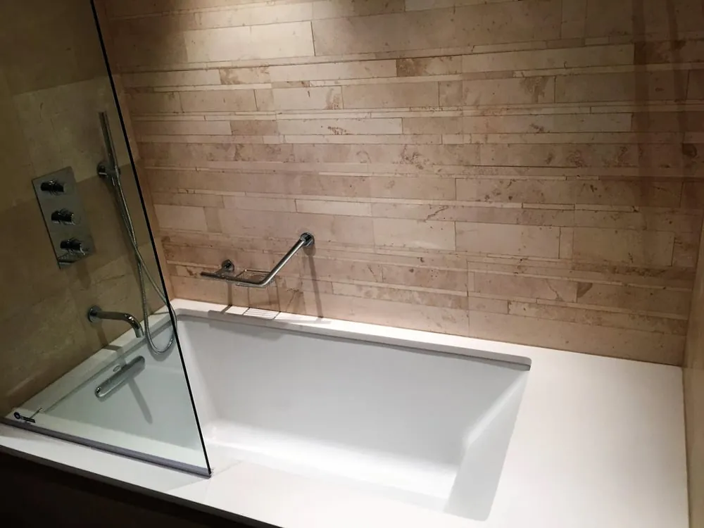 Deep drop-in soaking tub with fixed glass panel, beige limestone strip tile wall, chrome valve and hand shower.