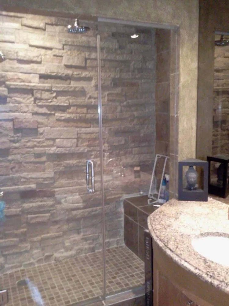 Glass-door shower with stacked stone wall, brown mosaic floor and tiled bench beside a granite vanity.