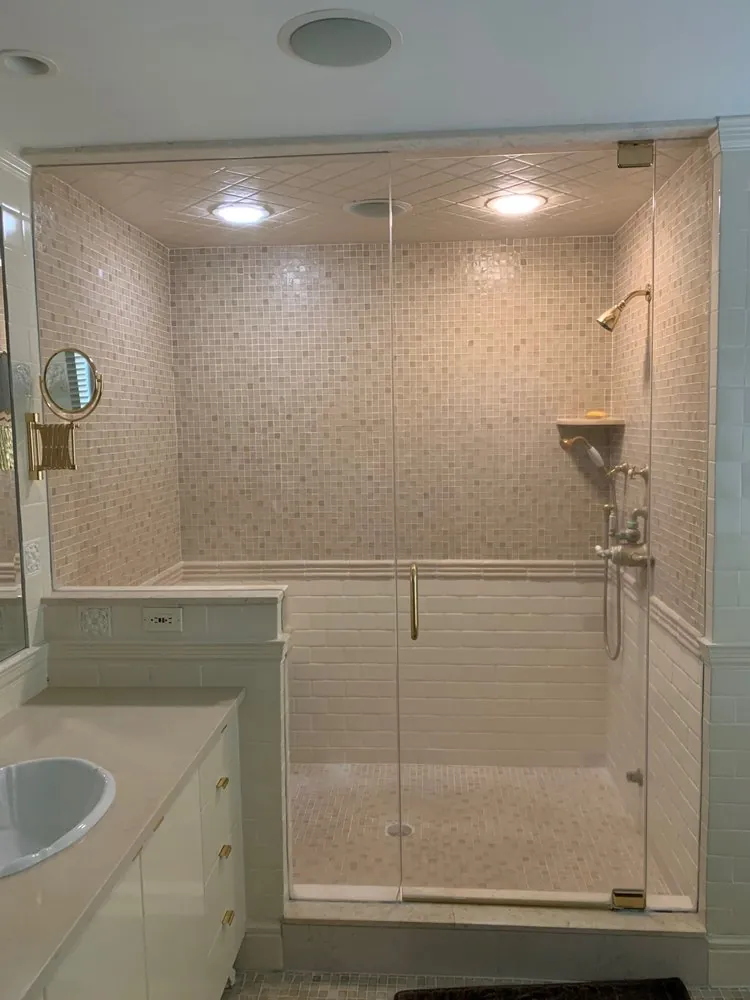 Large glass steam shower with beige mosaic tile, white subway wainscot, brass fixtures and a white vanity beside it.