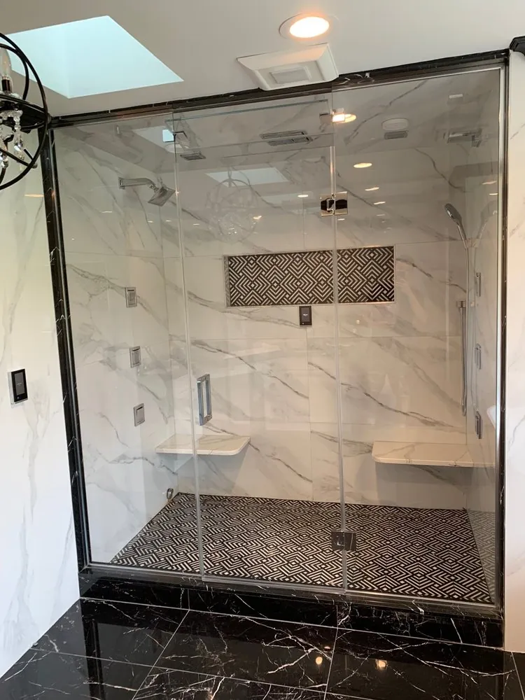 Large glass steam shower with white marble-look slabs, black and white geometric floor and niche, two benches, black floor.