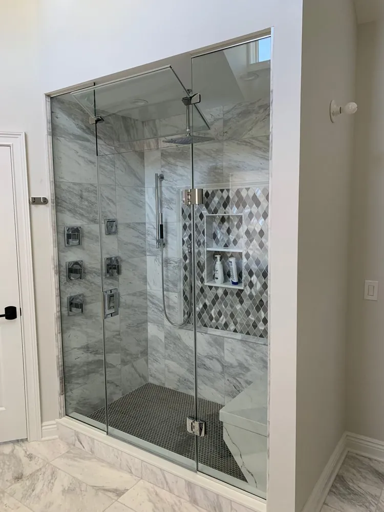 Wide view of a frameless glass steam shower with gray marble-look tile, body jets, bench and diamond mosaic niche.