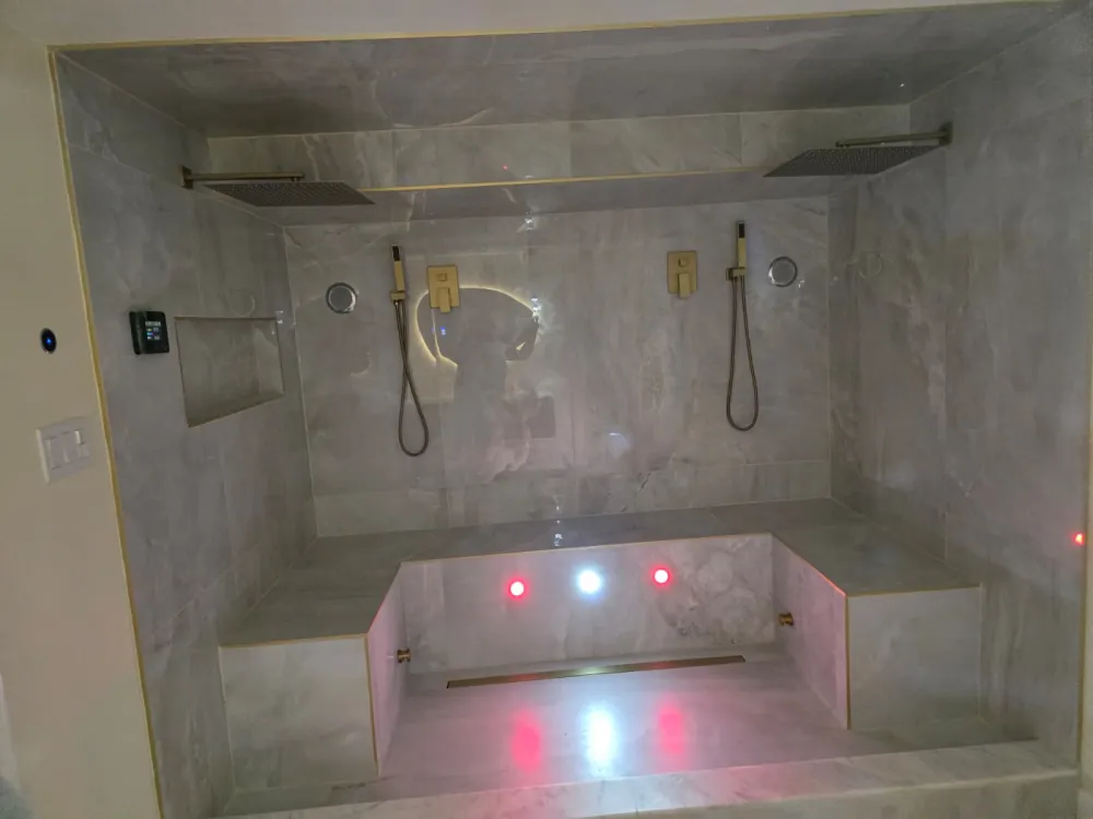 Double steam shower in light gray marble-look tile with two brushed gold rain heads, handhelds, a bench and colored lights.
